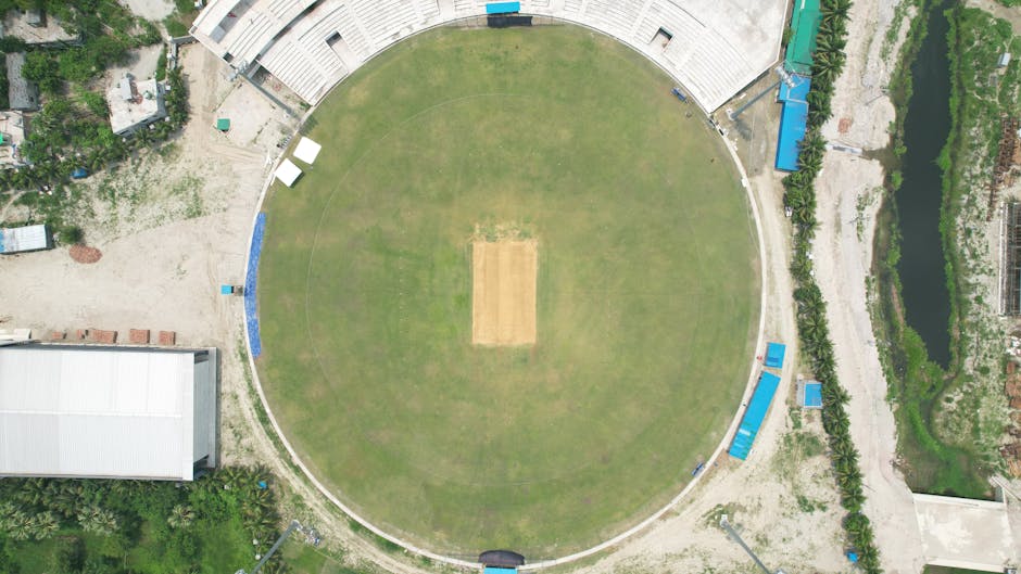 An aerial view of Lords Cricket Ground showing the oval-shaped pitch area in the center, surrounded by a green outfield with well-maintained grass. The seating stands are visible along the perimeter i