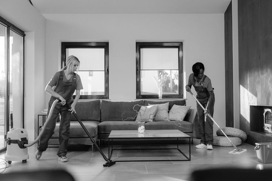 A woman with blonde hair, dressed in a grey t-shirt and red overalls, is standing in a modern kitchen cleaning the floor with a mop. The kitchen features white marble walls, a built-in oven, and woode