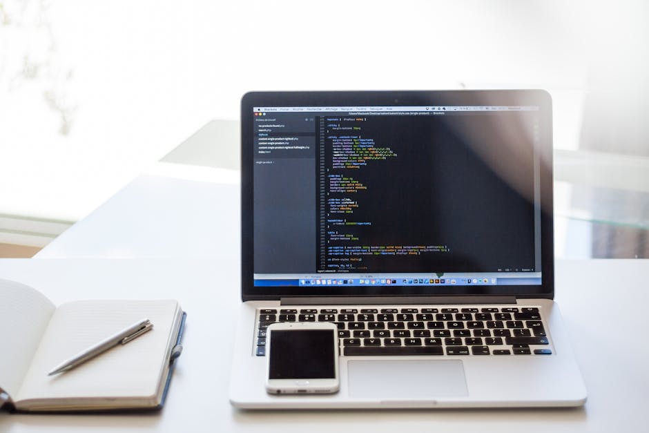 A close-up view of a silver laptop on a white desk, displaying lines of colorful computer code on its screen. Next to the laptop, there is a white smartphone placed flat on the desk and an open notebo